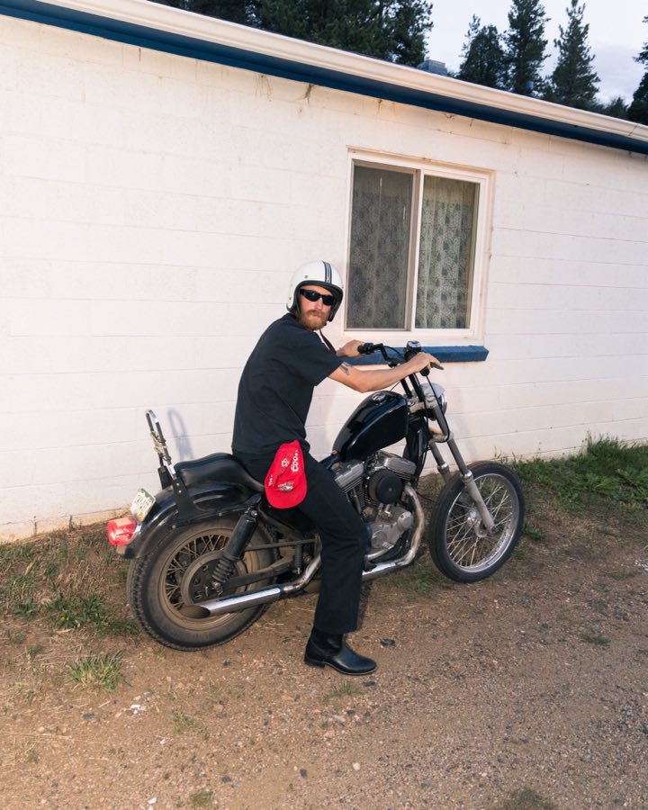 A rider in a white open-face helmet sitting on a black motorcycle beside a white cinder-block building, pine trees behind.